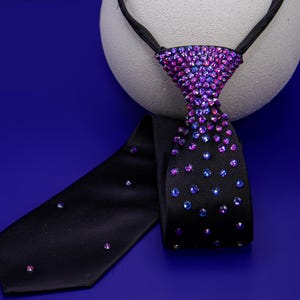 May include: A black necktie featuring a dense cluster of iridescent purple and blue rhinestones at the knot, which gradually transition into a sparse, scattered pattern of individual crystals extending down the length of the fabric.