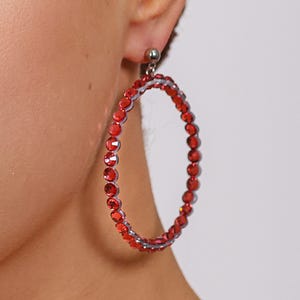 May include: A large circular hoop earring featuring a continuous row of vibrant red faceted crystals set in a metallic frame. The earring attaches to the earlobe with a small silver-toned ball stud post, creating a bold and reflective accessory.