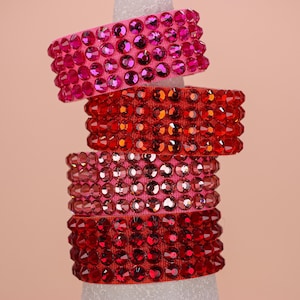 May include: A stack of four wide rings, each featuring four horizontal rows of faceted, sparkling crystals. Two rings are a vibrant hot pink, and two rings are a deep, saturated red. The crystals are set closely together on a fabric band.