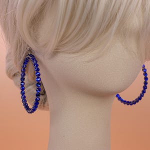May include: A pair of large hoop earrings featuring a circular frame wrapped in small, vibrant royal blue faceted beads. The beads are arranged in a continuous row around the entire circumference of each hoop, creating a textured, reflective surface.