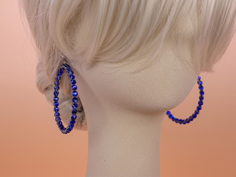 May include: A pair of large hoop earrings featuring a circular frame wrapped in small, vibrant royal blue faceted beads. The beads are arranged in a continuous row around the entire circumference of each hoop, creating a textured, reflective surface.