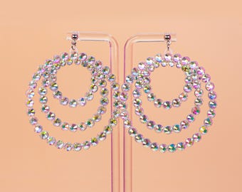 May include: A pair of iridescent hoop earrings. Each earring features three graduated circles of sparkling, multi-colored rhinestones. The earrings are suspended from a small silver-colored stud and are displayed against a peach-colored background.