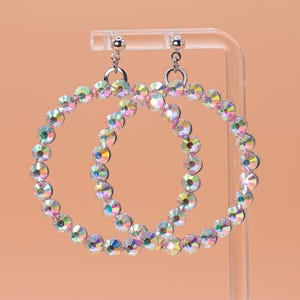 May include: A pair of large hoop earrings. Each earring is a circle of iridescent, multi-colored rhinestones. The earrings hang from silver-colored posts. The background is a solid, light orange color.