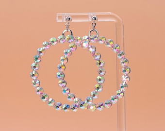 May include: A pair of circular hoop earrings featuring a continuous row of iridescent, multi-colored crystals set in a silver-toned metal frame. The earrings are suspended from small silver-toned ball studs, creating a reflective and shimmering effect.