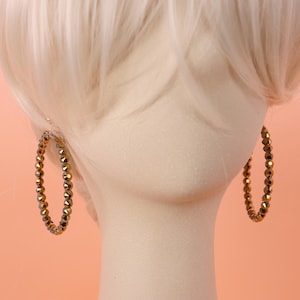 May include: A pair of hoop earrings featuring a continuous strand of small, faceted, metallic bronze-colored beads. The circular earrings are displayed on a neutral-toned mannequin head with light-colored hair, highlighting the reflective texture.