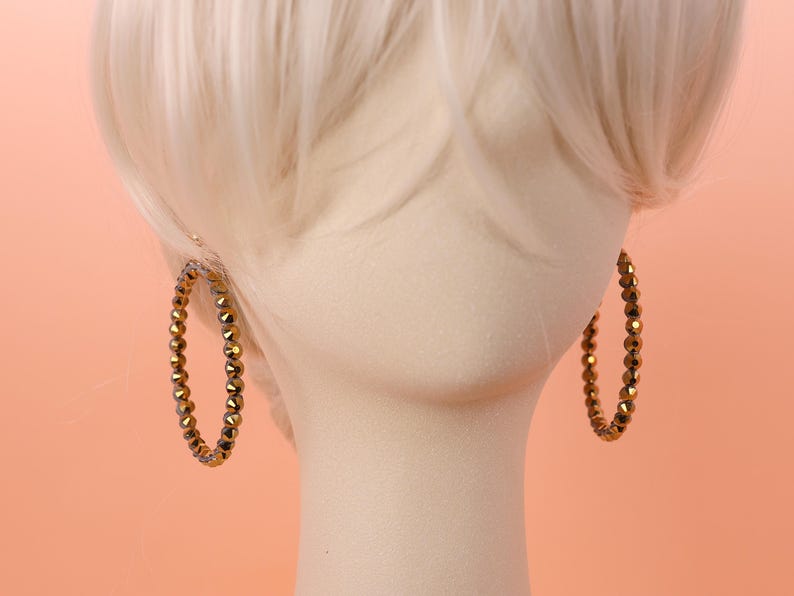 May include: A pair of hoop earrings featuring a continuous strand of small, faceted, metallic bronze-colored beads. The circular earrings are displayed on a neutral-toned mannequin head with light-colored hair, highlighting the reflective texture.