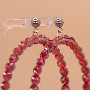 May include: A pair of hoop earrings featuring circular frames covered in faceted red crystals. The earrings are attached to silver-toned metal ball studs with small jump rings, creating a dangling design that catches the light.