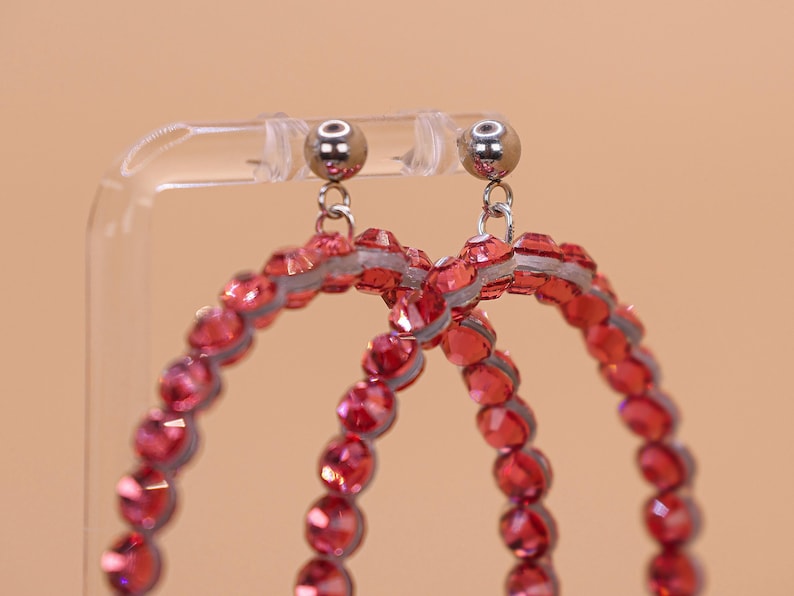 May include: A pair of hoop earrings featuring circular frames covered in faceted red crystals. The earrings are attached to silver-toned metal ball studs with small jump rings, creating a dangling design that catches the light.