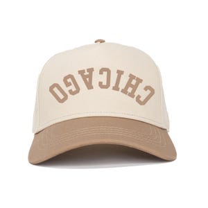 Könnte beinhalten: Beige-braune Baseballkappe mit dem Wort "CHICAGO" in geschwungenen Großbuchstaben. Die Kappe hat einen gebogenen Schirm und eine strukturierte Krone. Der Text ist in einer Blockschrift gehalten.