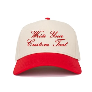 May include: A baseball cap with a cream-coloured crown and a red brim. The cap features the embroidered text "Write Your Custom Text" in red script. The cap has a red button on top.