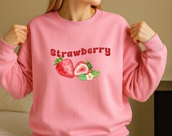 Puede incluir: Sudadera rosa claro con la palabra "Strawberry" en escritura roja. El gráfico presenta una fresa entera y una fresa cortada, junto con hojas verdes y flores blancas. La sudadera es de color rosa liso.
