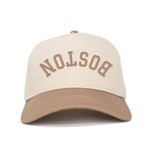Op de afbeelding: Een beige en bruine baseballcap met het woord "BOSTON" in verhoogde blokletters op de voorkant. De cap heeft een gebogen klep en een mesh achterkant. De hoed is neutraal van kleur en geschikt voor dagelijks gebruik.