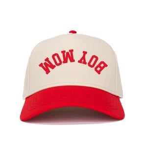 May include: A baseball cap with a cream-coloured crown and a red brim. The text "BOY MOM" is printed in red across the front. The cap has a curved brim and a small red button on top.