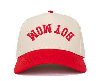 Pode incluir: Boné de beisebol com coroa creme e aba vermelha. O texto "BOY MOM" está impresso em vermelho na frente do boné. O boné tem uma aba curva e um pequeno botão vermelho no topo.