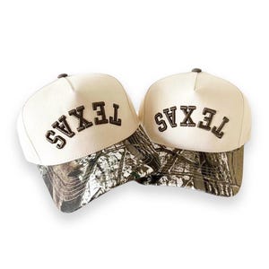 Puede incluir: Dos gorras de béisbol color crema con la palabra "TEXAS" bordada en marrón en la parte delantera. La visera de cada gorra presenta un estampado de camuflaje en tonos marrones, verdes y grises. Las gorras se muestran sobre un fondo blanco.