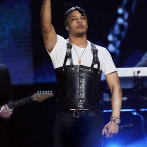 May include: A performer on stage wearing a white t-shirt, black leather corset with gold-colored rivets, and black pants. The performer is holding a microphone and wearing a watch and necklace.