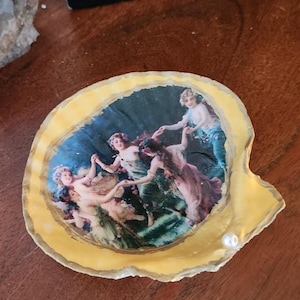 May include: A yellow seashell with a gold trim, decorated with a vintage image of five women dancing in a circle. The image is set against a dark blue background with green foliage.