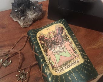 Può includere: Un diario rilegato in pelle verde con un'illustrazione della carta dei tarocchi de "Gli Amanti" è esposto. Il diario è allacciato con un cordoncino di cuoio marrone. È visibile anche un cordoncino di cuoio marrone con un ciondolo a forma di timone e un ciondolo a forma di ancora.