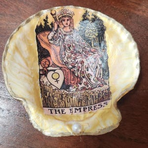 May include: A golden seashell with a pearl in the centre. The shell features a tarot card image of The Empress, a woman seated on a throne with a crown and a sceptre. The card is surrounded by a golden border and the text "THE EMPRESS" is printed below the image.