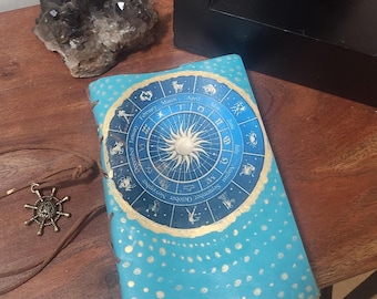 Puede incluir: Un diario de cuero azul con un diseño de rueda zodiacal dorada y lunares blancos. La rueda zodiacal presenta los meses del año y los signos astrológicos.