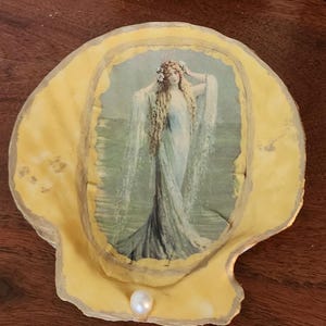 Puede incluir: Una concha de mar amarilla con un borde dorado, una perla y una imagen vintage de una mujer con el pelo largo de pie en el océano.