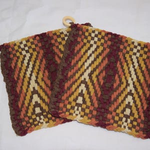 May include: Two woven potholders with a geometric pattern in shades of brown, orange, yellow, and cream. Each potholder has a wooden ring for hanging. The design features a diamond and chevron pattern.