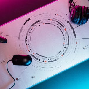 May include: A large rectangular desk mat with a light gray surface featuring a central circular geometric pattern in black, gray, and orange. A black wired computer mouse, a black game controller, and black over-ear headphones rest on the mat.