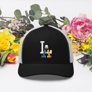 Könnte beinhalten: Eine schwarze und weiße Trucker-Kappe mit einem weißen "LA" bestickt auf der Vorderseite. Das "A" besteht aus einer Regenbogen-Pride-Flagge.
