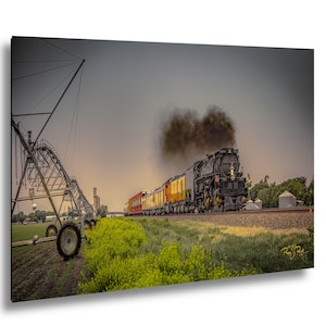 May include: A black steam locomotive pulling a train with yellow and orange cars travels along tracks next to a green field with a large metal irrigation system. Dark smoke billows from the engine into a grey sky above the rural landscape.