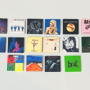 Może przedstawiać: Wystawa na ścianie 15 małych kwadratowych płócien, każde z inną okładką albumu. Okładki obejmują zespoły takie jak The Beatles, Nirvana, Queen, The Smiths i AC/DC. Płótna są ułożone w siatkę.
