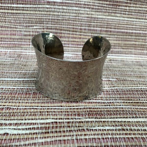 May include: A wide, silver-colored cuff bracelet with a textured, hammered metal finish. The bracelet has a curved shape and an open design. The jewelry is displayed on a woven, neutral-toned surface.