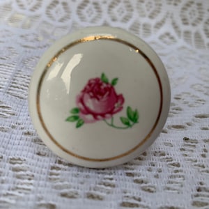 May include: A round, off-white ceramic cabinet knob featuring a central pink rose illustration with green leaves. The knob is accented with a thin, circular gold-colored rim around the edge, providing a polished finish to the floral design.