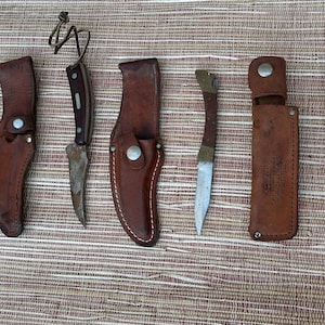 May include: A collection of seven vintage knives and a sharpening steel. The knives have brown leather sheaths and metal blades. The sharpening steel is made of metal and has a wooden handle.