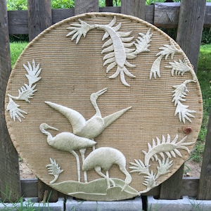 May include: Round woven wall art featuring a beige background with white crane figures and plant-like accents. The cranes are in various poses, with one in flight. The artwork is displayed outdoors against a wooden fence.