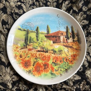 May include: White ceramic plate with a painted landscape scene. The artwork depicts a field of sunflowers, a house, and cypress trees under a blue sky. The plate has a decorative border and a vintage aesthetic.