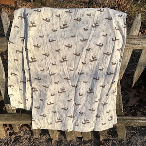 May include: A white fabric with a repeating pattern of brown ducks swimming in a pond with green reeds. The fabric is draped over a wooden fence.