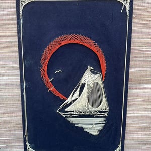 May include: A navy blue string art piece featuring a sailboat with white thread, set against a red sun. The artwork includes a white border and two small birds. The sailboat is reflected in the water.