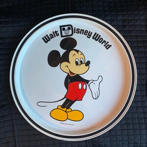 May include: A white, round Walt Disney World serving tray featuring a smiling Mickey Mouse cartoon character. The tray has a black and white border and the words "Walt Disney World" in black text.