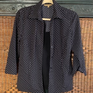 May include: Black and white polka dot button-up shirt with a long sleeve. The shirt has a collar and is hanging on a wooden hanger.