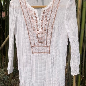 May include: White long-sleeve tunic with a V-neck and intricate brown embroidery. The embroidery features floral and geometric patterns. The fabric has a textured, pleated appearance. The tunic is hanging on a wooden hanger against a bamboo backdrop.