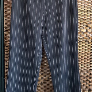 May include: Black pinstripe trousers hanging on a black hanger. The pants have a straight leg cut and a classic pinstripe pattern with thin white stripes. The trousers are displayed against a woven background.