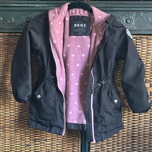 May include: Black hooded jacket with a pink fleece lining featuring a white heart pattern. The jacket has a zipper closure, two pockets, and a drawstring at the waist. The DKNY logo is visible on the front of the jacket.