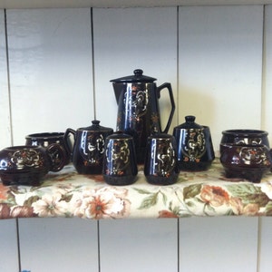 May include: A set of brown ceramic kitchenware with black and gold floral designs. The set includes a coffee pot, creamer, sugar bowl, and four salt and pepper shakers.