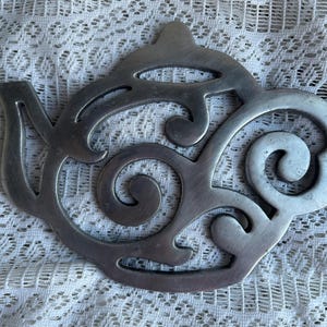 Puede incluir: Un salvamanteles de metal plateado con forma de tetera. El salvamanteles tiene un diseño decorativo calado con patrones arremolinados. Está diseñado para proteger las superficies de los utensilios de cocina calientes.