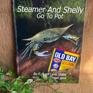 May include: A book titled "Steamer And Shelly Go To Pot" featuring a crab illustration and an Old Bay Seasoning can. The book cover is dark with a crab and bubbles. The Old Bay can is yellow, red, and blue.