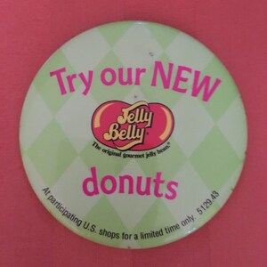 May include: A round button with a light green diamond pattern background. The button features the text "Try our NEW" in pink, the Jelly Belly logo, and the word "donuts" in pink. The text "At participating U.S. shops for a limited time only" is also present.