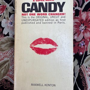 May include: A vintage paperback book cover with the title "CANDY" in bold, black letters. The cover is white with a red lipstick print and the text "GOOD GRIEF! THIS IS THE ORIGINAL FAMOUS BOOK - THAT'S COMING SOON AS A SUPER MOVIE" and "NOT ONE WORD CHANGED! This is the ORIGINAL, UNCUT and UNEXPURGATED edition as first published and banned in Paris." The author's name, "MAXWELL KENTON", is printed at the bottom of the cover.