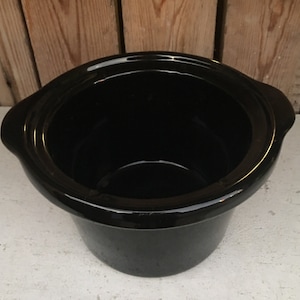 May include: A black, oval-shaped slow cooker insert with a glossy finish. The insert has two handles on either side and a wide opening. The interior is smooth and dark, contrasting with the lighter surface it rests on.
