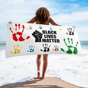 May include: White beach towel with colorful handprints and the text "BLACK LIVES MATTER" in black. The handprints are in red, yellow, green, blue, and brown. The towel is held up by a person on a sandy beach with the ocean in the background.