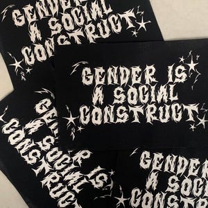 May include: Close-up of four black fabric patches with white text and star designs. The text on the patches reads "Gender is a social construct".